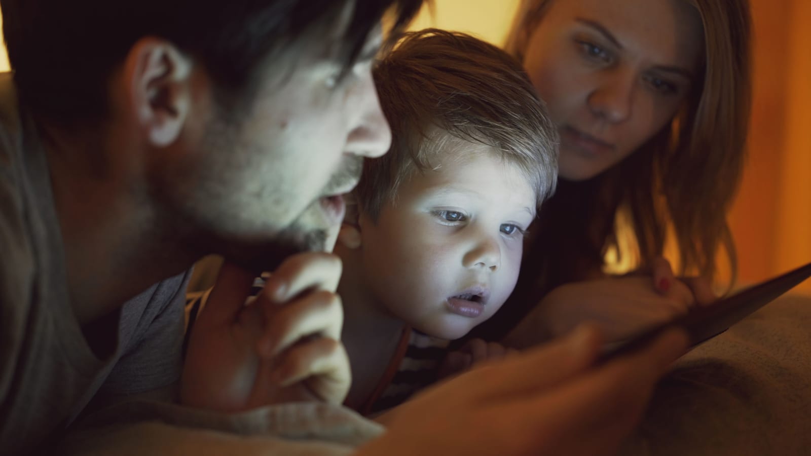 Rodiče a malé dítě v noci společně zírají do tabletu, dětský obličej ozářený displejem