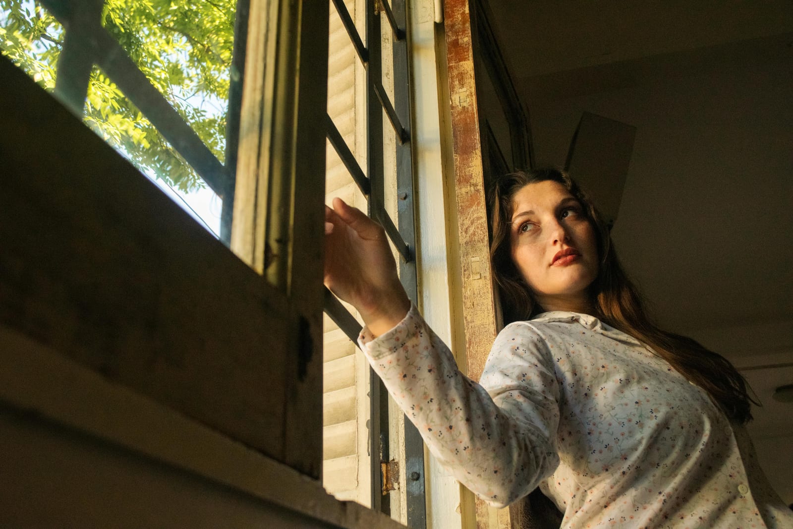 Mladá žena u otevřeného okna, ruka opřená o rám, hledí zamyšleně ven do koruny stromu