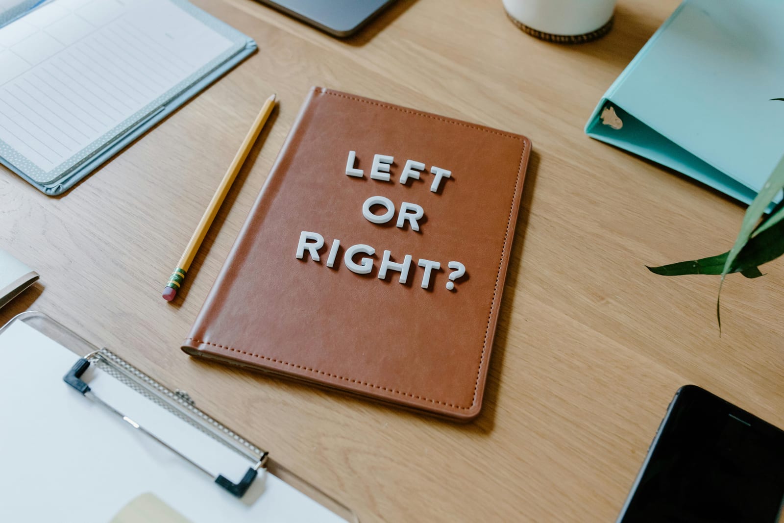 Pohled shora na dřevěný stůl s hnědým notesem, na kterém jsou bílá písmena LEFT OR RIGHT; kolem notes, propiska, plánovač a klipboard — moment rozhodování