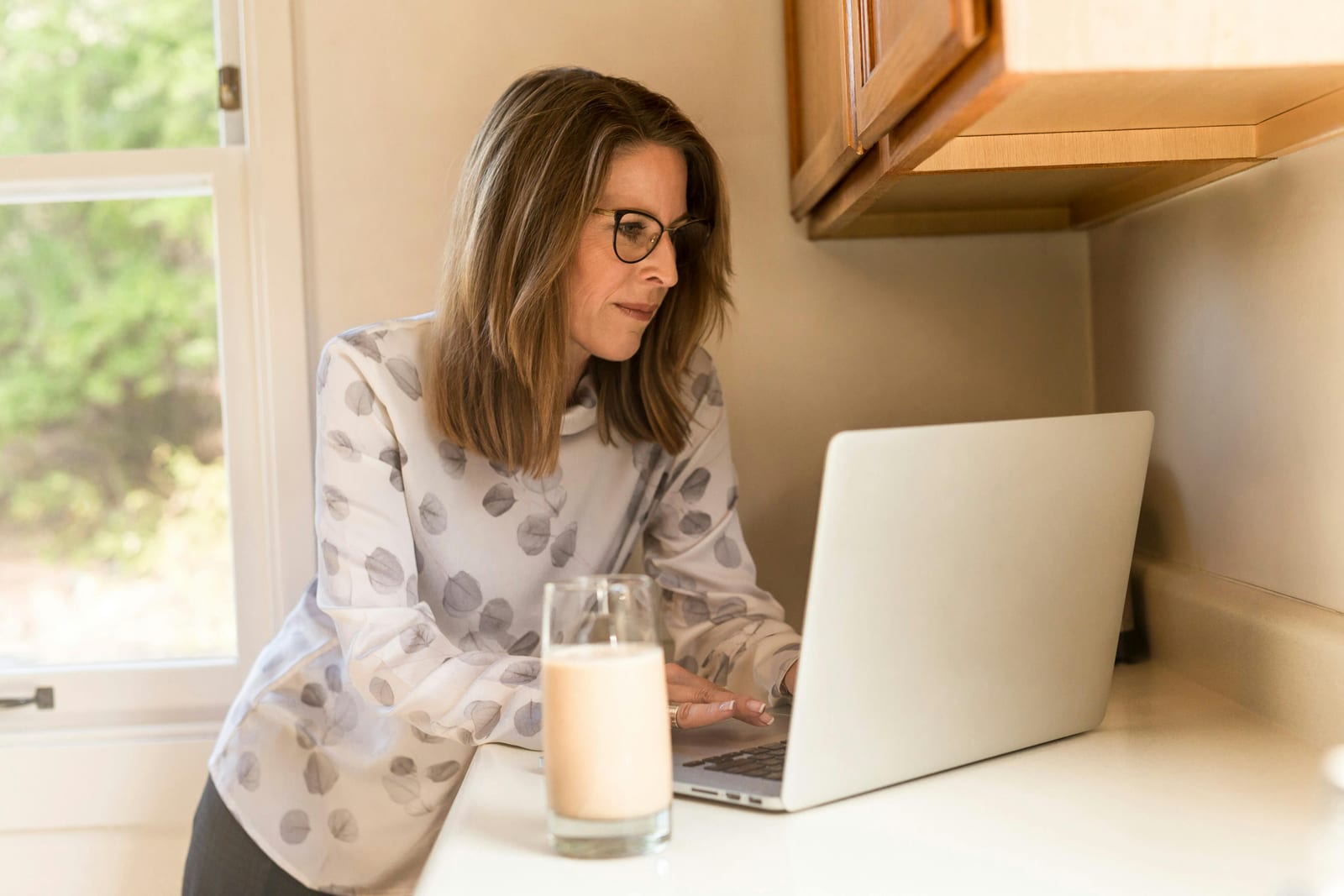 Žena s brýlemi se naklání nad notebookem u kuchyňské linky, vedle sklenice se smoothie a otevřené okno do zahrady