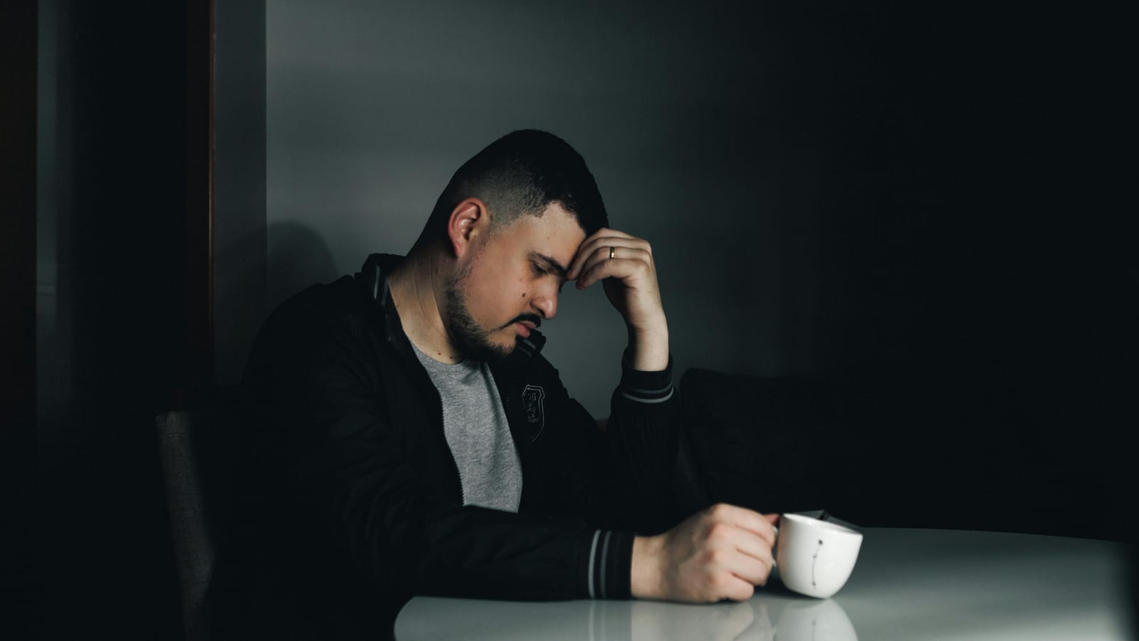 Mladý muž v tmavé bundě sedí osamoceně u stolu, čelo si opírá o ruku, před sebou má bílou kávovou misku, tlumené světlo dopadá z bočního okna na zamyšlený výraz