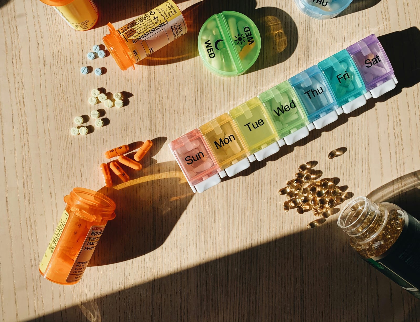 Týdenní dávkovač léků s barevnými oddíly na všechny dny, kolem rozsypané pilulky a oranžové lékovky