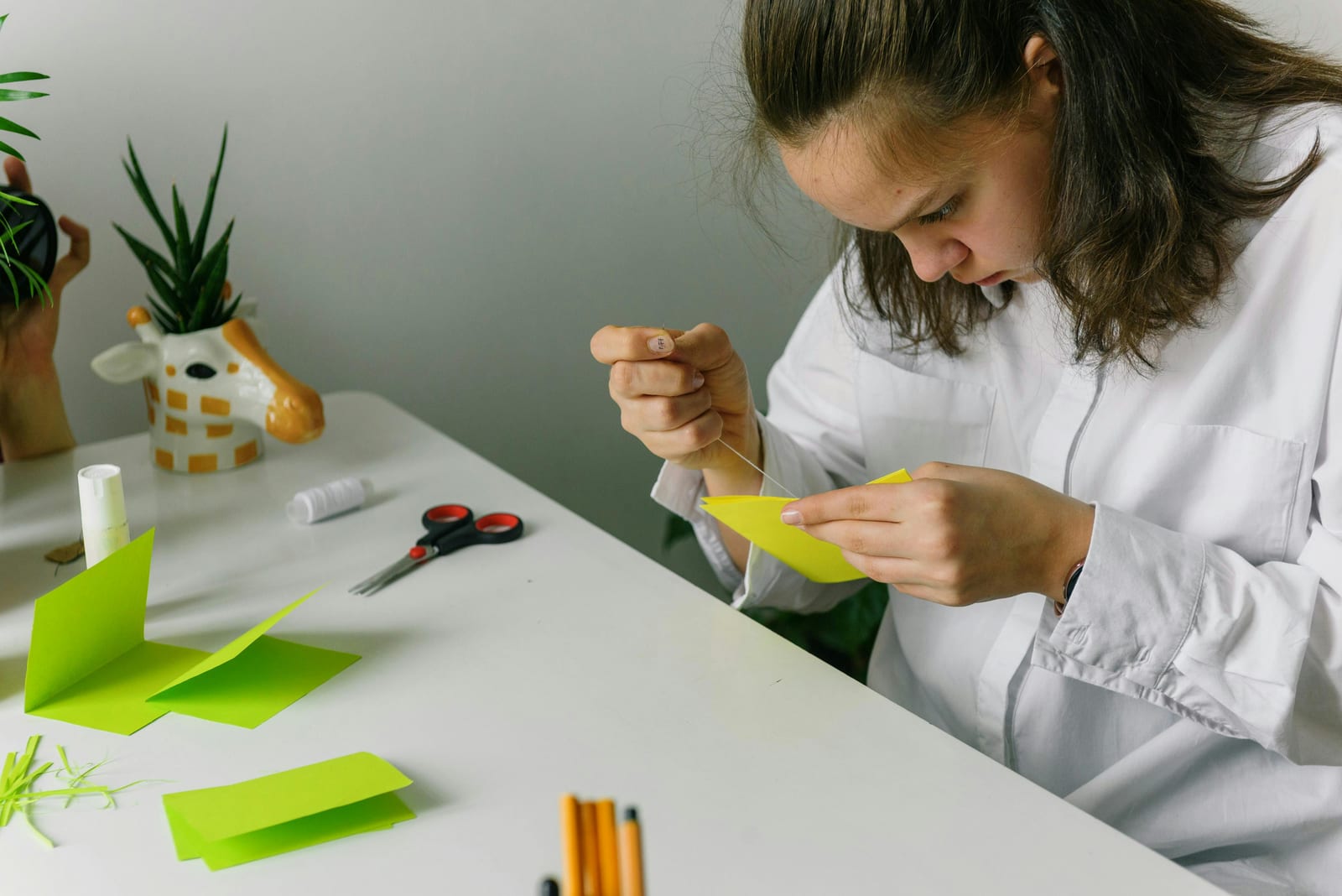 Žena v bílé košili soustředěně tvoří origami u stolu se zelenými papíry, nůžkami a fixy