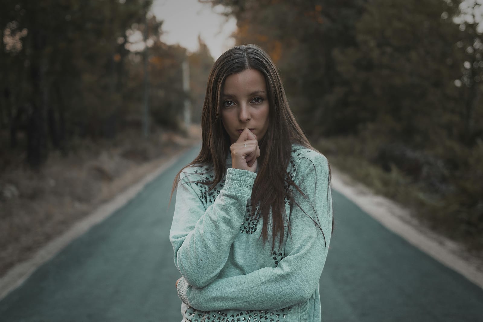Mladá žena stojí uprostřed lesní cesty, ruku u brady, a zamyšleně hledí před sebe