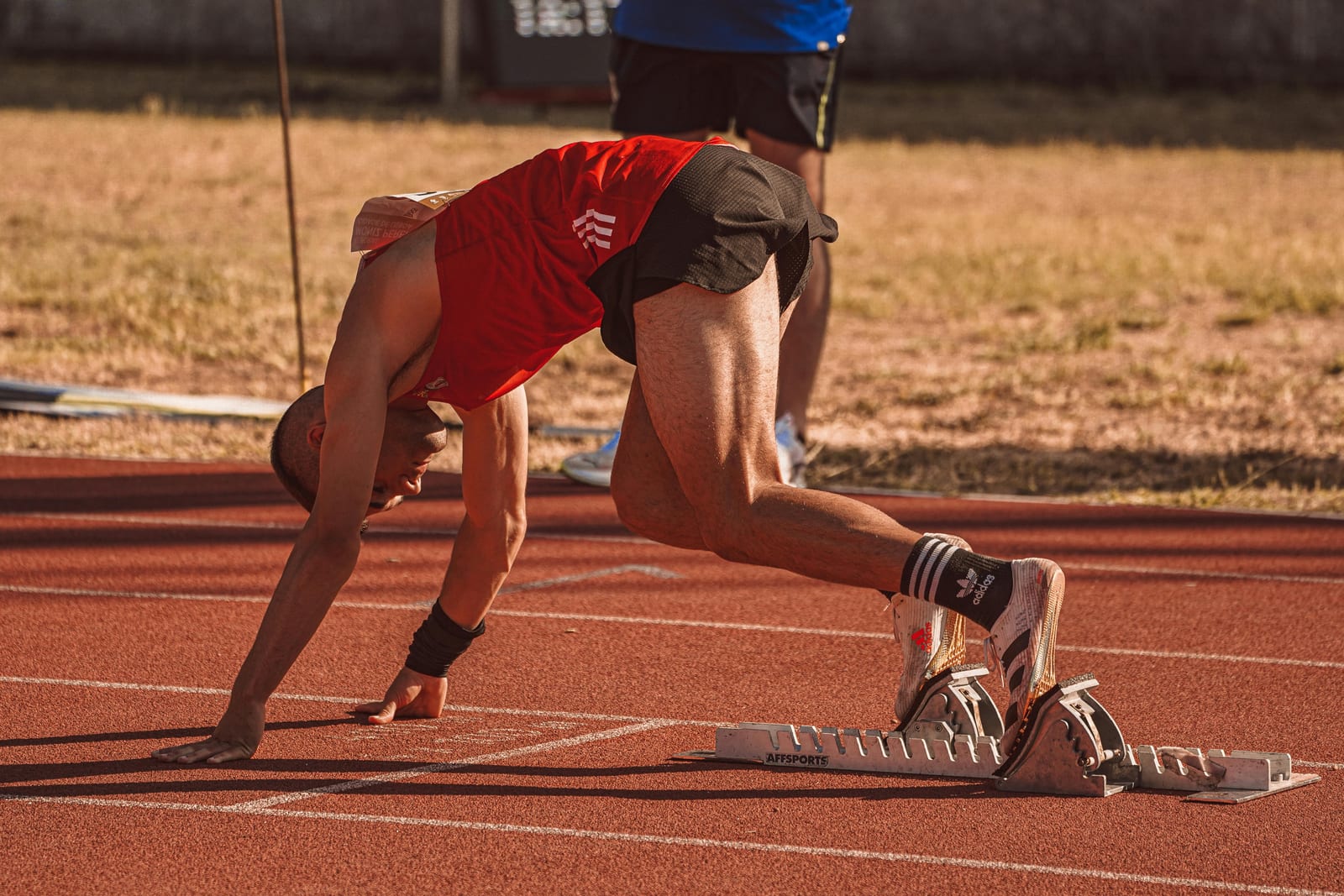 Sprinter v červeném dresu v nízkém startu na atletické dráze, soustředění před prvním krokem, sluneční světlo