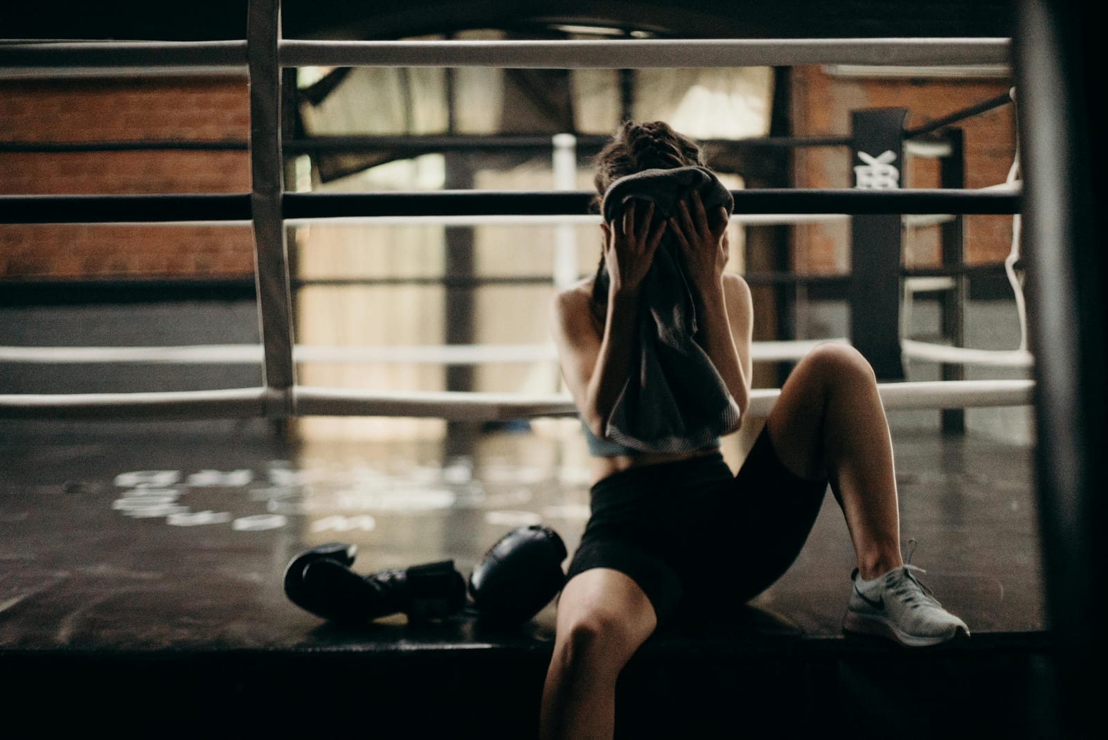 Žena v sportovním oblečení sedí na okraji boxerského ringu a otírá si obličej ručníkem po tréninku