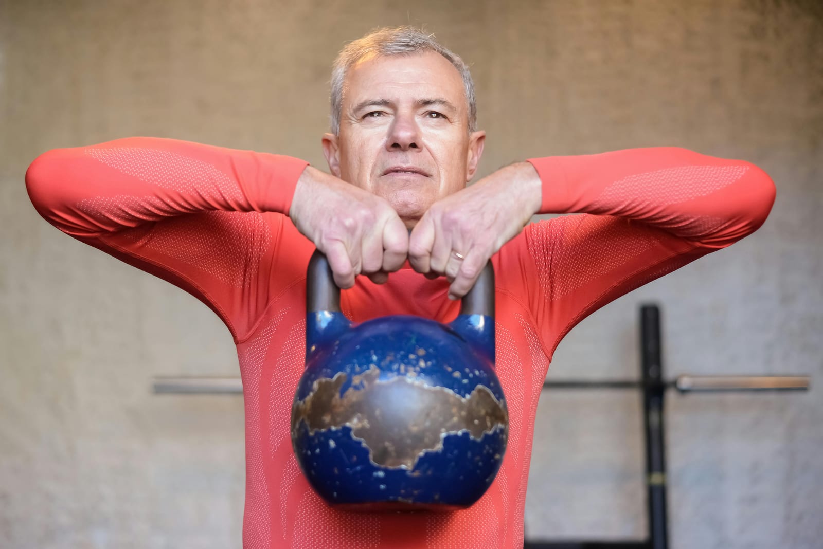 Starší muž v červeném sportovním tričku drží v rukou před hrudí těžký kettlebell