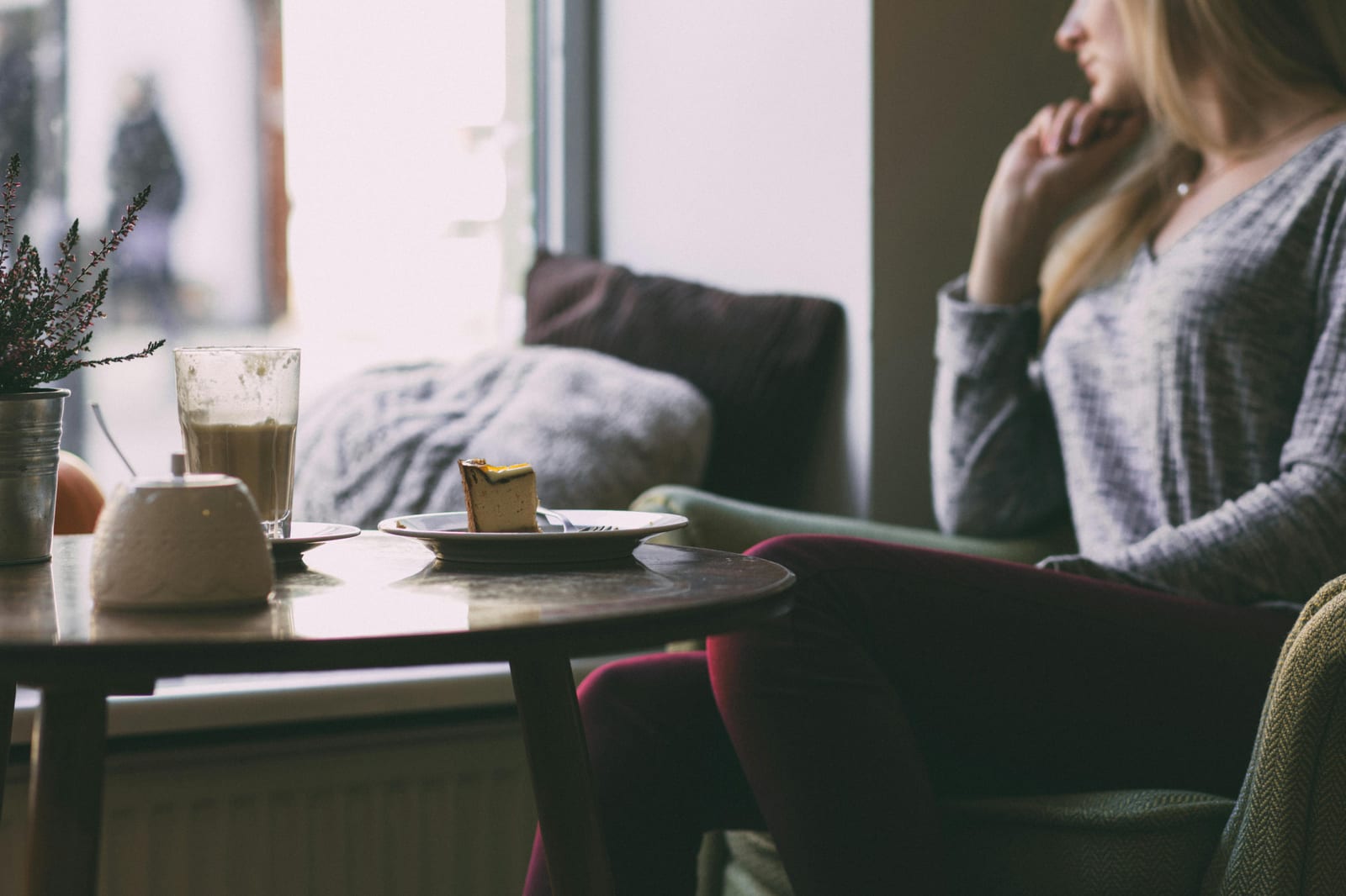 Žena sedí v kavárně u okna s šálkem latté a kouskem dortu na stole, prsty u brady — okamžik pauzy mezi rozhodnutími