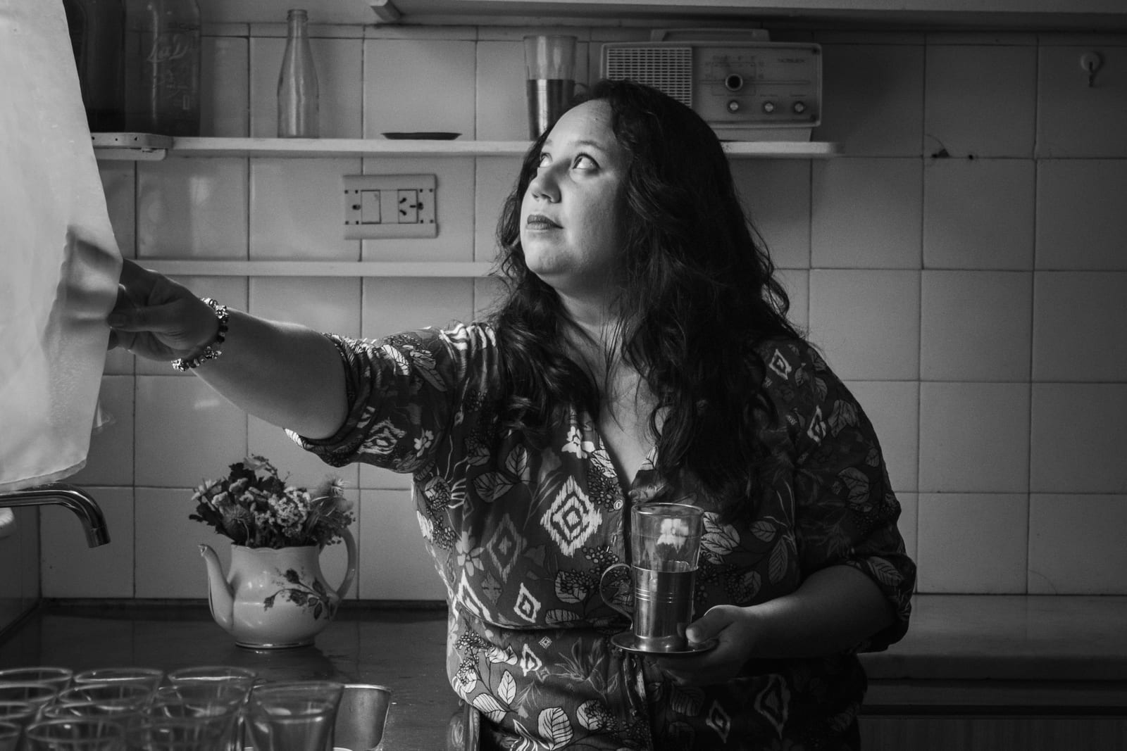 Černobílá fotografie ženy v květovaných šatech stojící v kuchyni; jednou rukou se opírá o závěs, v druhé drží šálek a hledí mimo objektiv s odevzdaným, zamyšleným výrazem