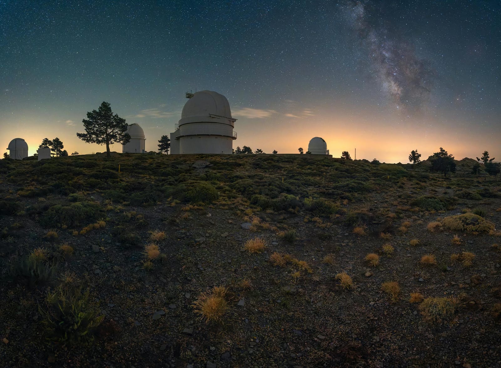 Astronomická observatoř s několika kupolemi na vrcholku kopce pod hvězdnou oblohou s viditelnou Mléčnou dráhou