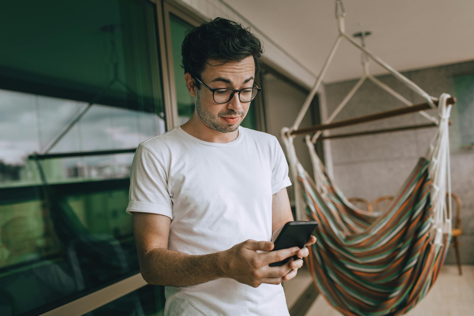 Muž s brýlemi v bílém tričku stojí na balkoně a s nejistým výrazem se dívá do mobilu